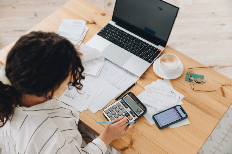 Top View Of A Woman Working At Home In The Kitchen With Financial Papers, Counting On A Calculator, Paying Bills, Planning A Budget To Save Some Money. Independent Accounting, Remote Accountant